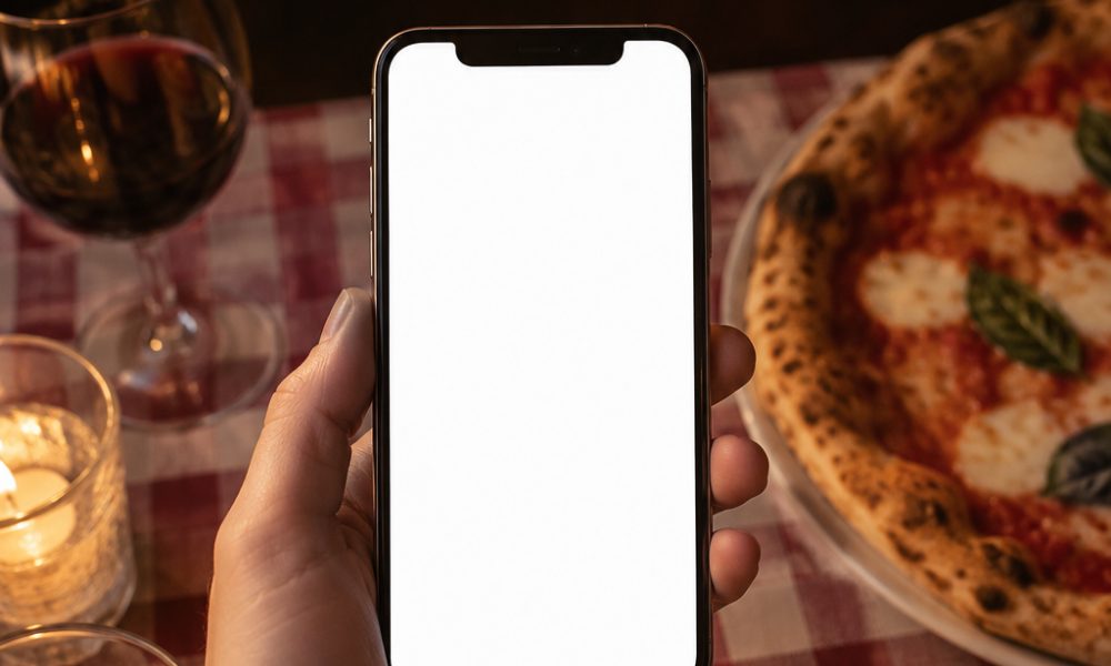 Un cliente con il telefono in mano al tavolo di un ristorante, accanto a una pizza e a un calice di vino
